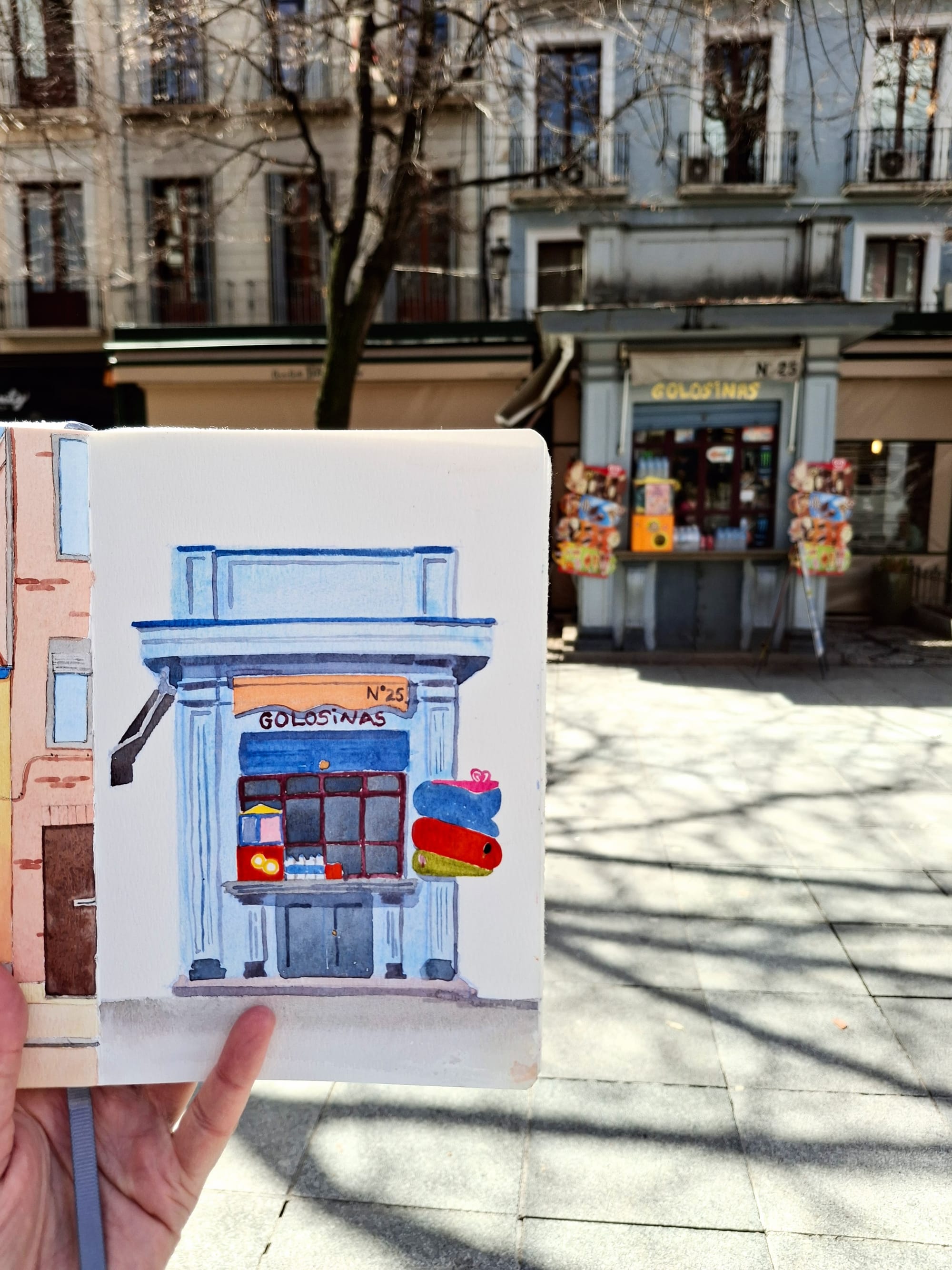 A hand holds a watercolour sketch of a blue kiosk called "Golosinas" with a candy machine and colourful signage. In the background, the real kiosk is visible.