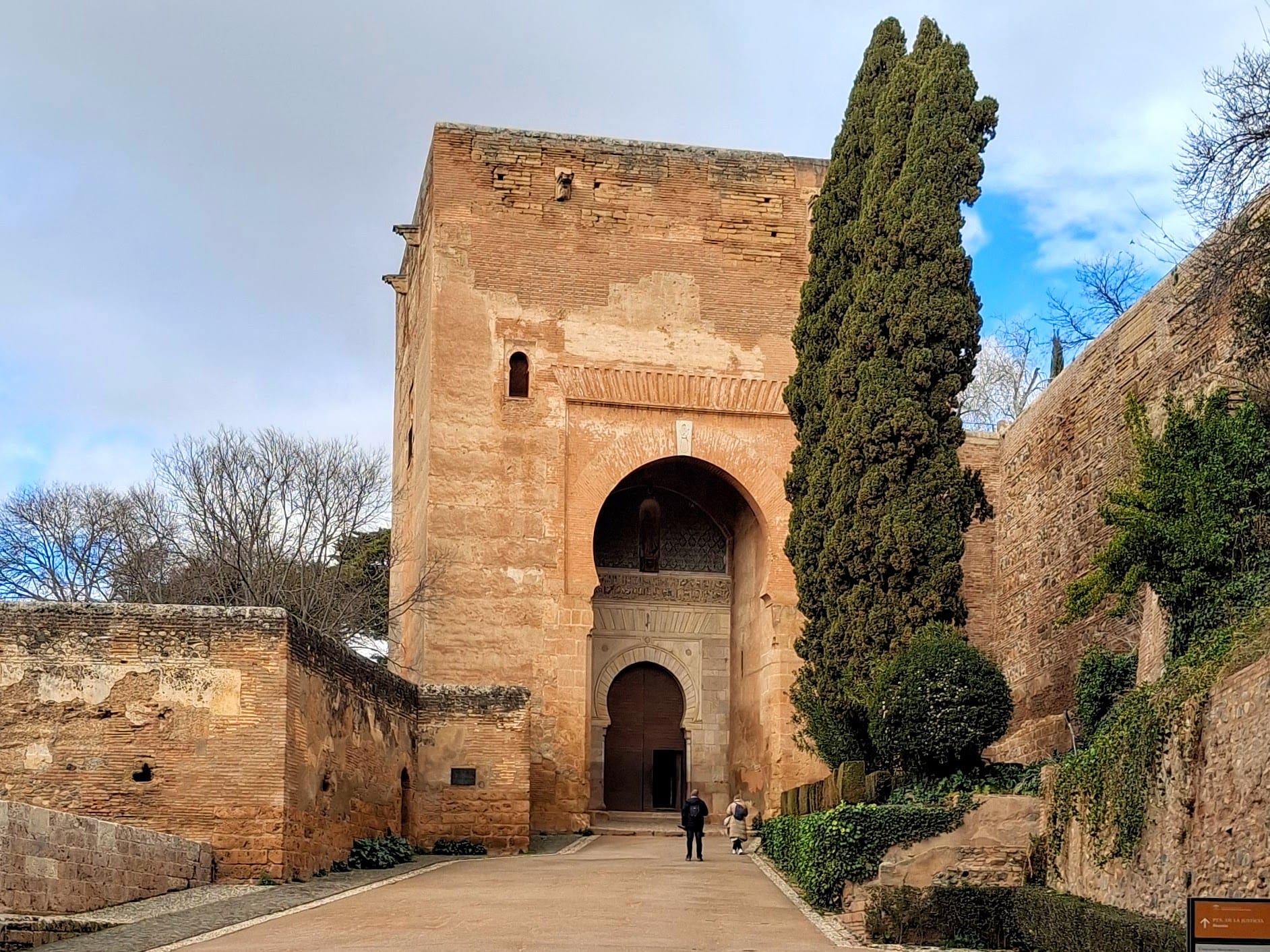 Photo of a large stone gate in sandy beige. Two tiny people stand in front of the gate, which is framed by greenery and stone walls. The sky is cloudy and some winter trees are visible in the distance.