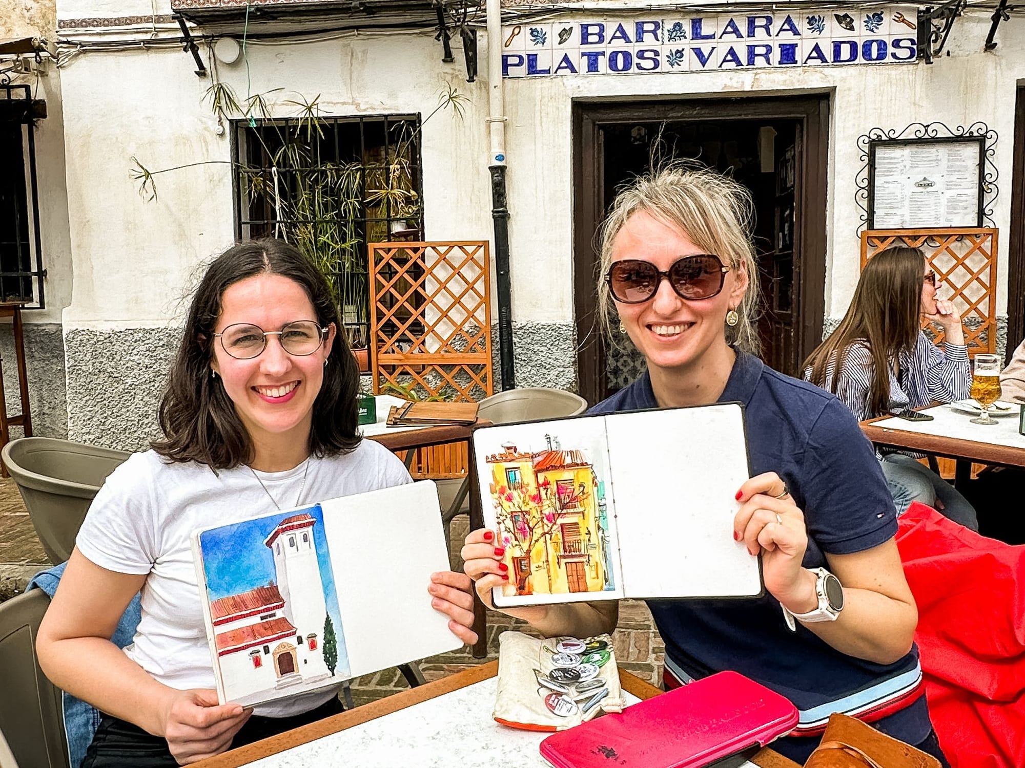 Two white women sitting in a coffee shop in the Albaicín in Granada and holding up watercolour sketches with big smiles. 