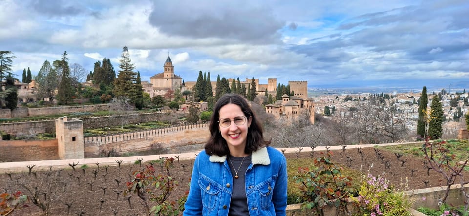 A white woman is a blue denim jacket smiling at the camera in front of the old palace at the Alhambra, Granada on a cloudy day.